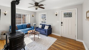 A living room with a black wood-burning stove, a blue sofa, a ceiling fan, and a white door with a decorative glass panel. - Red Door Cottage | 1/2 Mile to Whiskey Row (Prescott)