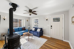 A living room with a black wood-burning stove, a blue sofa, a ceiling fan, and a white door with a decorative glass panel. - Red Door Cottage | 1/2 Mile to Whiskey Row (Prescott)