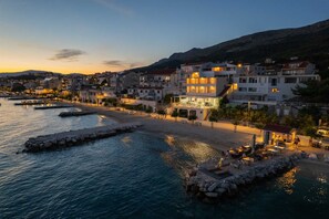 A coastal town at dusk with buildings, a beach, and a pier. - Beachfront Villa Victoris – Private Sunbeds (Podstrana)