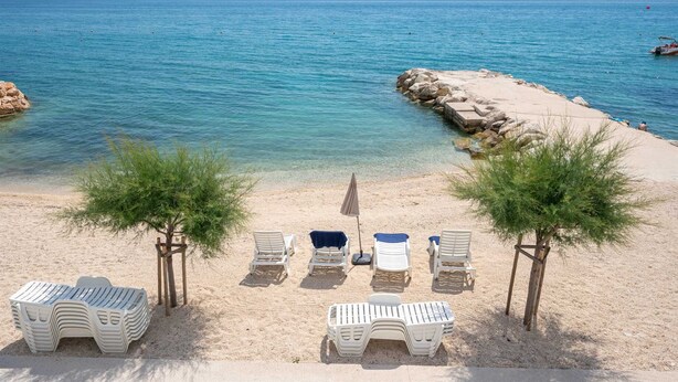 A beach with clear blue water, a sandy shore, and white lounge chairs arranged for sunbathing. - Beachfront Villa Victoris – Private Sunbeds (Podstrana)