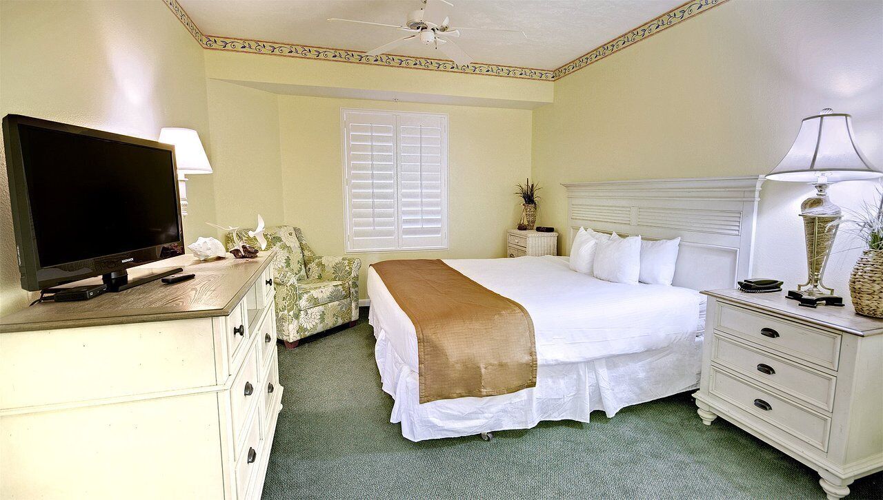 Habitación de hotel con cama, televisión, escritorio, silla y ventana con persianas.