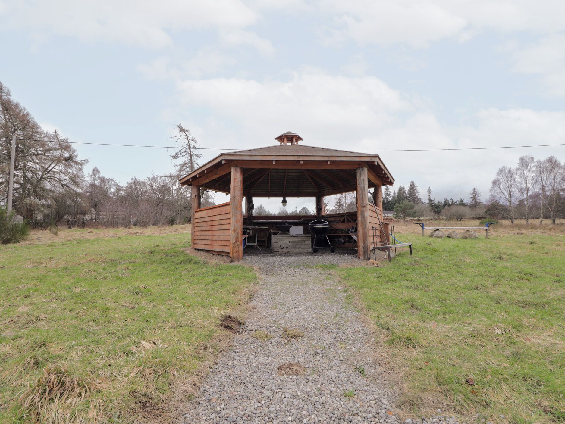 A wooden gazebo with a gravel path leading to it, surrounded by grass and trees.