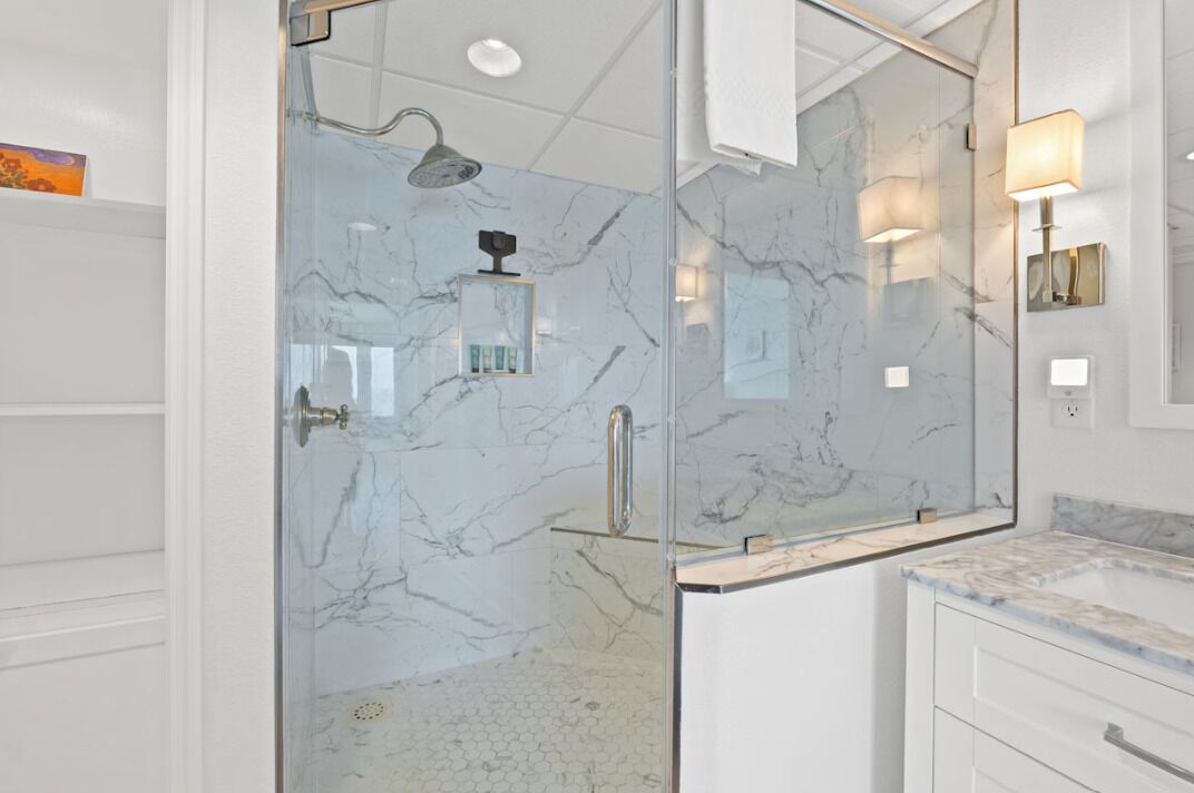 Une salle de bain moderne dotée d’une douche à parois de verre, de murs en marbre et d’un meuble-lavabo blanc avec un plan de travail en marbre.