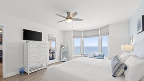 A modern bedroom with a large bed, a ceiling fan, a TV, and a view of the ocean. - Emerald Beach 1736 (Panama City Beach)