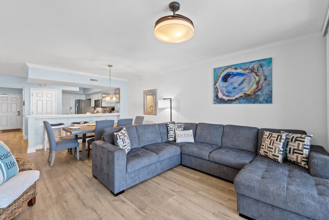 A modern living room with a grey sectional sofa, a dining area with a white table and chairs, and a kitchen in the background.