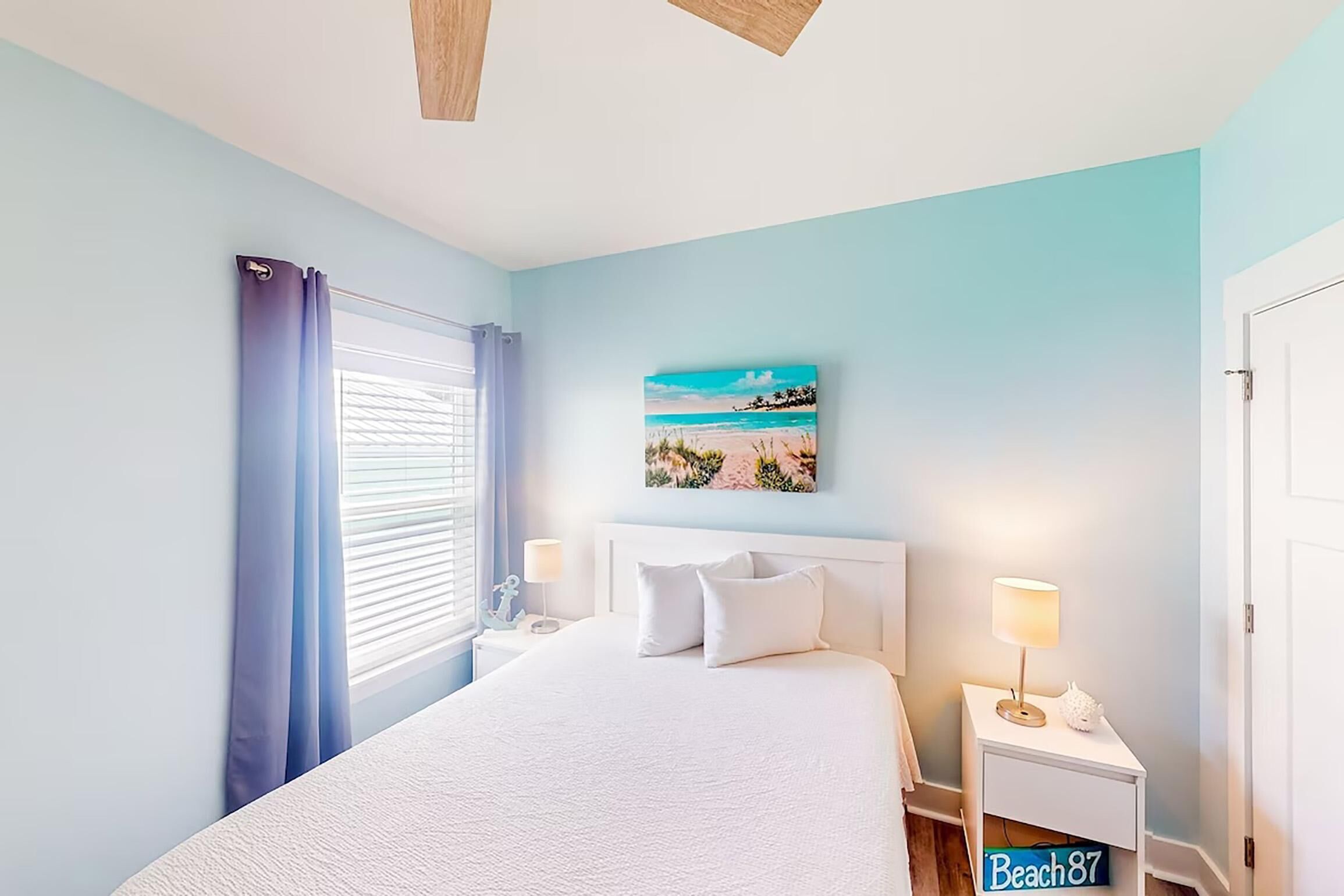 A bedroom with a bed, two bedside tables with lamps, a window with curtains, and a beach-themed wall art.