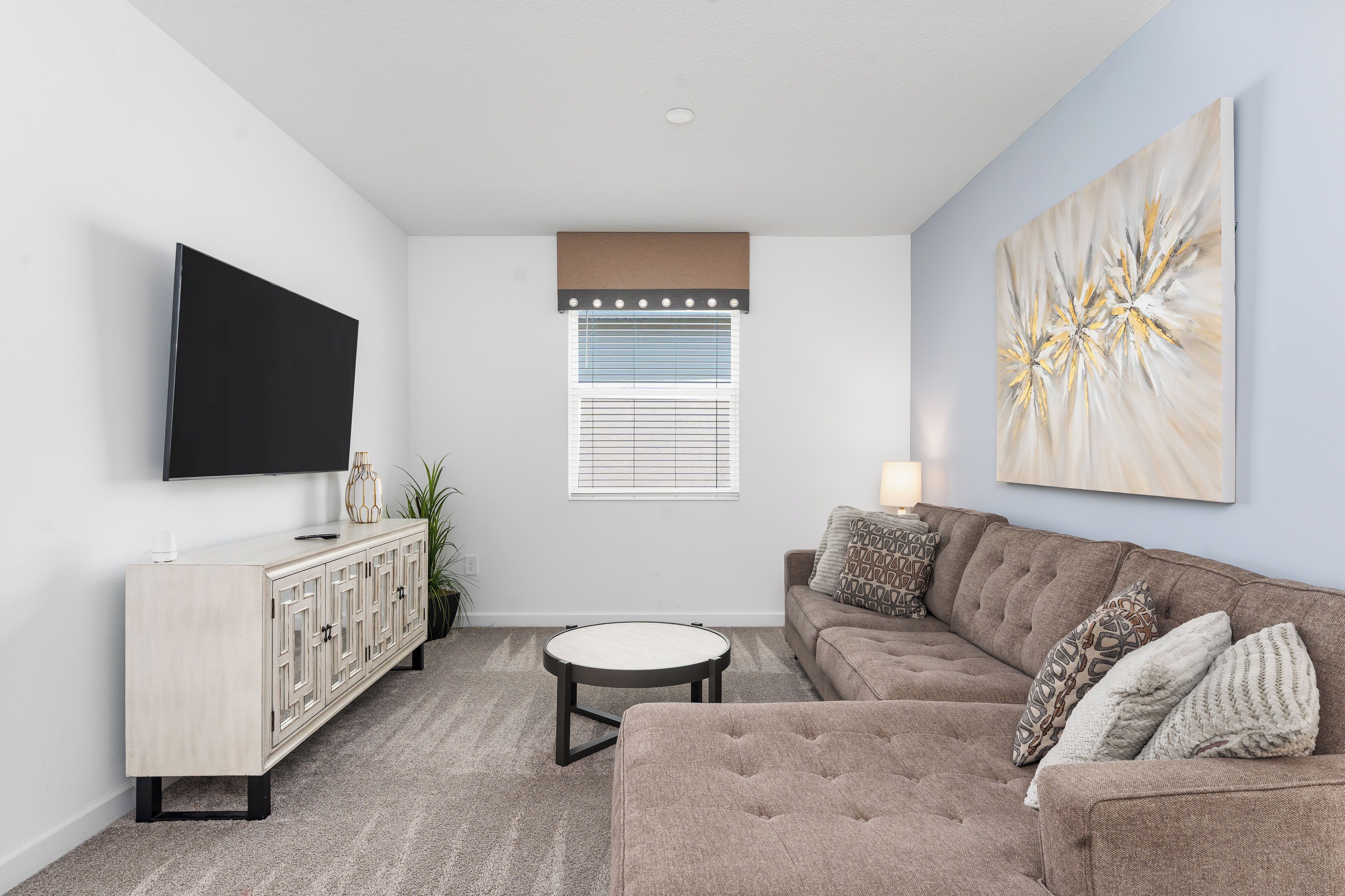A living room with a flat-screen TV, a sofa, a round coffee table, a cabinet, and a window with blinds.