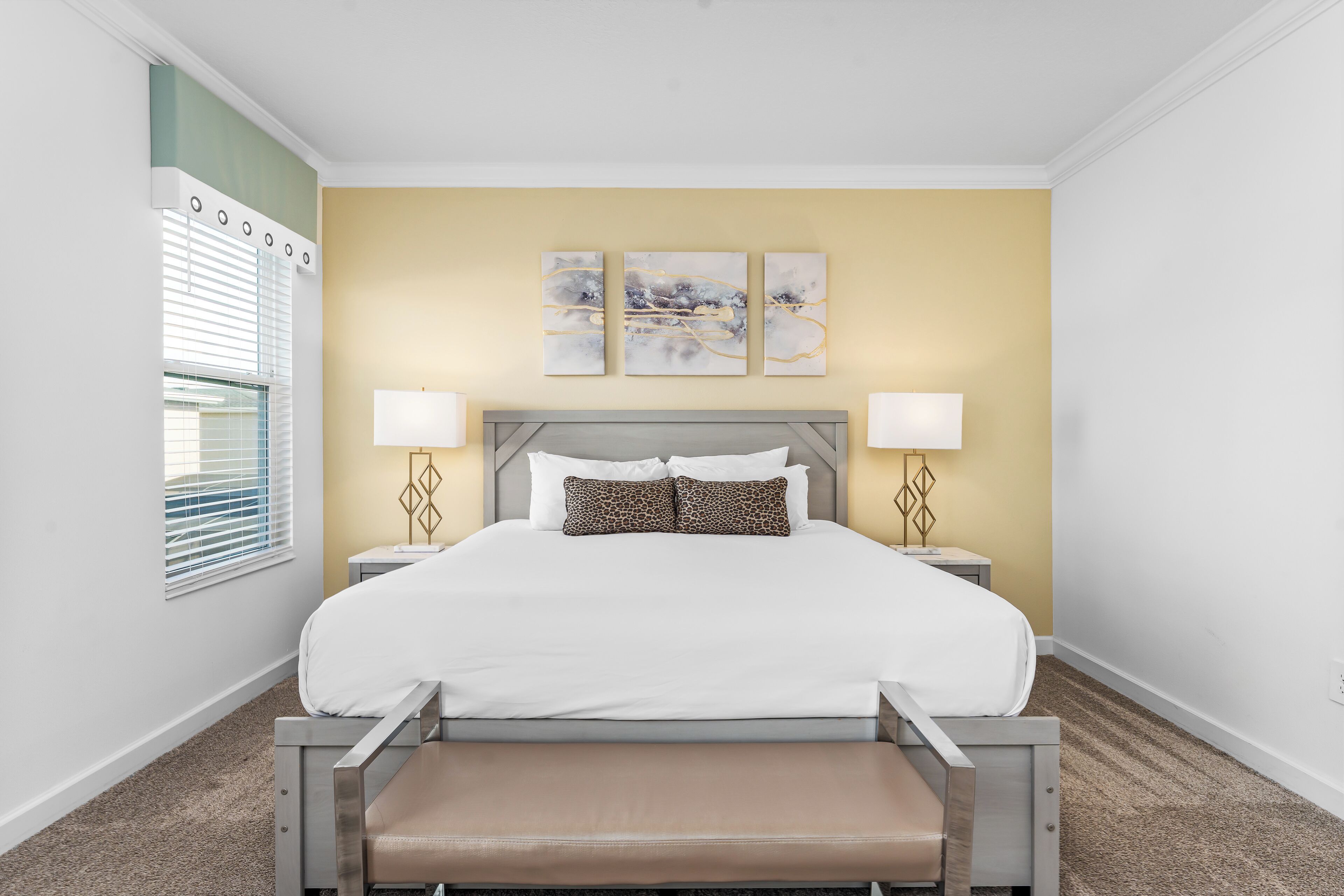 A bedroom with a bed, bedside tables, lamps, a bench, and artwork on the wall.