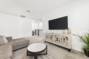 A living room with a sofa, a round coffee table, a TV on a white wall, a cabinet with decorative items, and a potted plant. - Kids Bedrooms | Game Area | Screened Private Pool (Kissimmee)