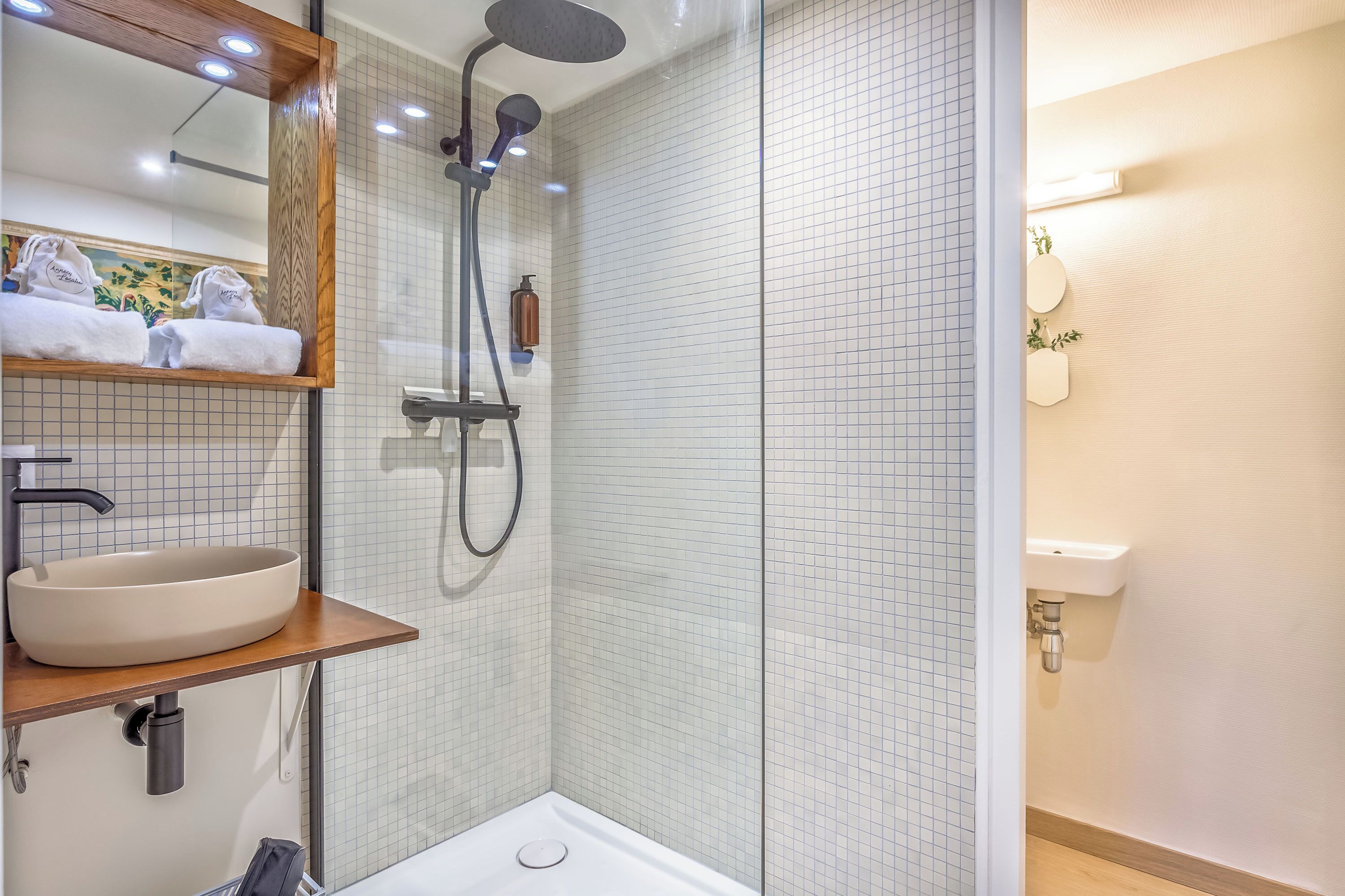 A modern bathroom with a large mirror, a round sink, a shower with a handheld showerhead, and a towel rack with folded towels.