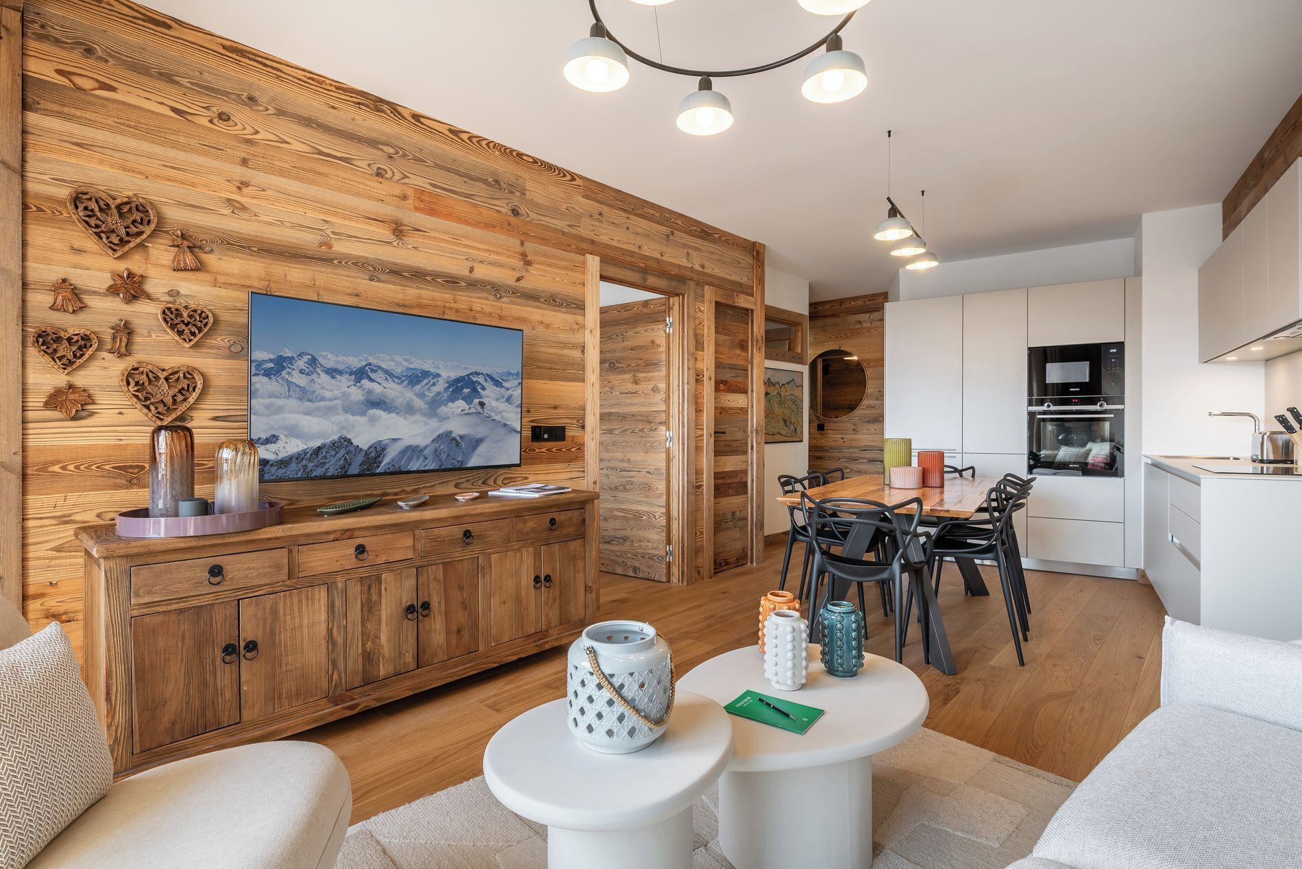 A modern living room with wooden paneling, a large TV, and a dining area with a kitchen in the background.
