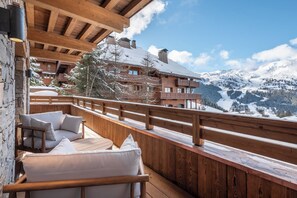 A wooden deck with outdoor seating, overlooking a snow-covered mountain landscape and a chalet-style building.
