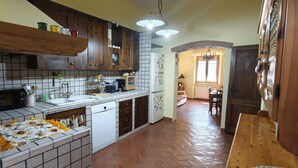 Une cuisine avec des meubles en bois, un évier blanc et un plan de travail carrelé. On y trouve un coin repas avec une table et des chaises, ainsi qu’une fenêtre avec des rideaux.