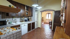 Eine Küche mit Holdekor-Schränken, einem weißen Spülbecken und einer gefliesten Spritzschutzwand. Es gibt einen Essbereich mit Tisch und Stühlen, sowie ein Fenster mit Vorhängen.