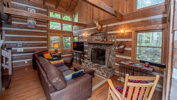 A cozy cabin interior with a stone fireplace, leather sofa, and large windows offering views of the forest. - 3 Brothers Rustic Sabbatical Creekside Cabin (Walland)