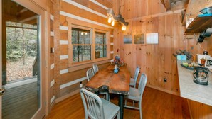 A wooden cabin interior with a dining table, chairs, a chandelier, and a view of the forest through large windows. - 3 Brothers Rustic Sabbatical Creekside Cabin (Walland)