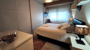 Een moderne hotelkamer met een groot bed, een nachtkastje met een lamp, een klein bureau met een schaaltje snacks, en een raam met gordijnen.