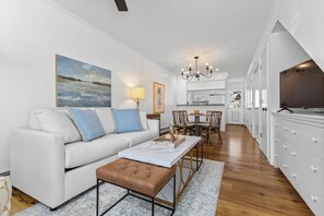 Una sala de estar moderna con un sofá blanco, un reposapiés marrón y una mesa de centro de mármol. Hay una zona de comedor con una mesa y sillas, y una cocina al fondo.