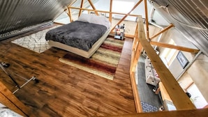 A loft bedroom with a wooden floor, a bed with a dark blanket, and a view of the sky through a skylight.