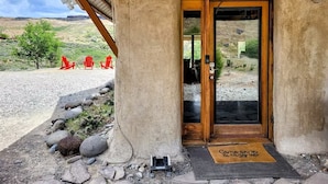 A rustic building with a wooden door and a welcome mat. Outside, there are red chairs and a gravel path.