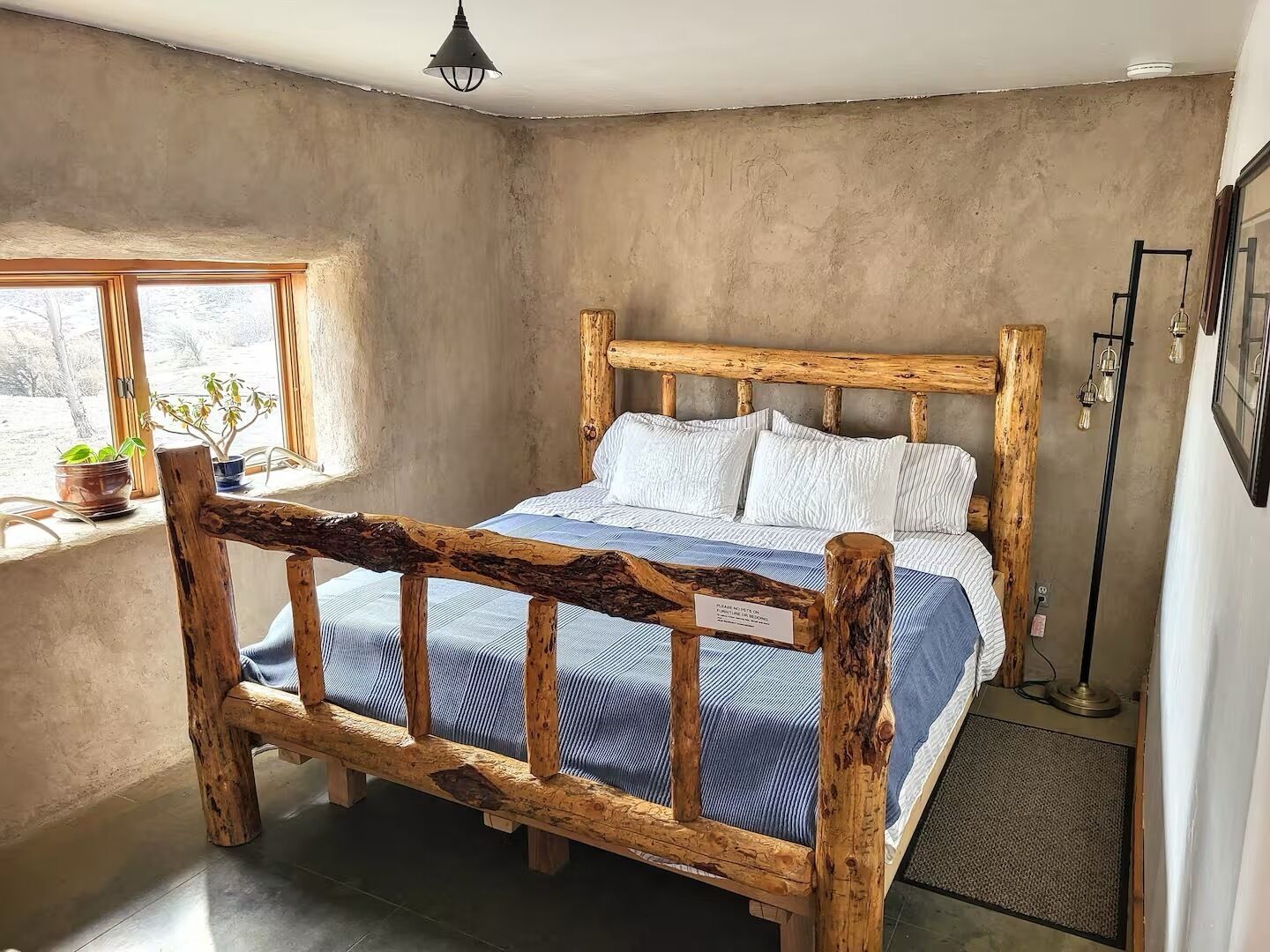 A rustic bedroom with a wooden bed frame, a window with plants, and a bedside lamp.