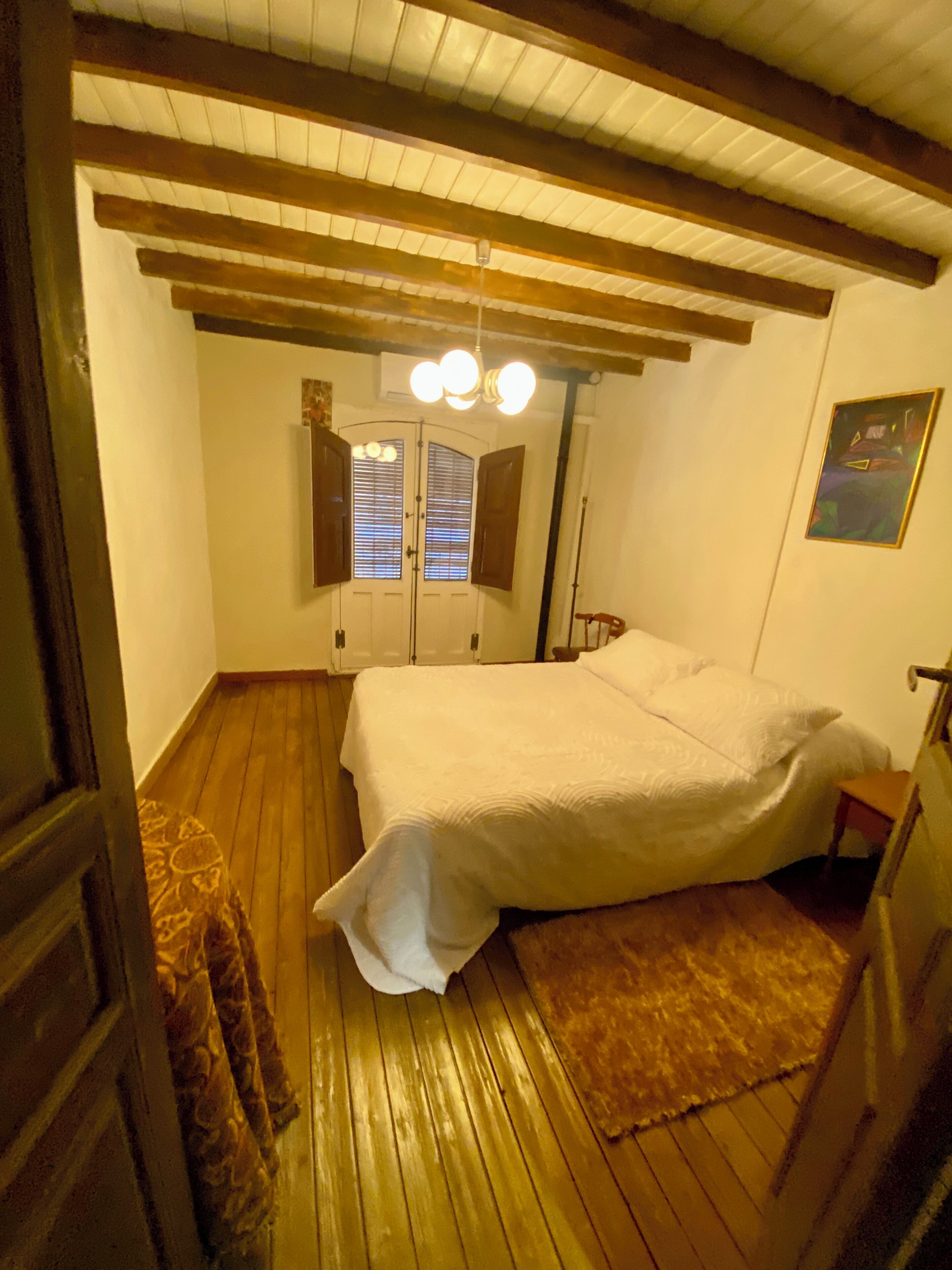 A bedroom with a wooden floor, a bed with white bedding, a small wooden chair, and a ceiling light fixture.