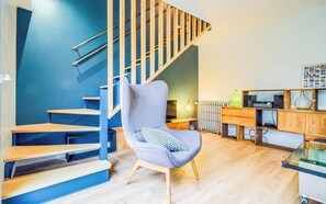 A modern living room with a blue wall, wooden staircase, a grey armchair, and a glass coffee table.