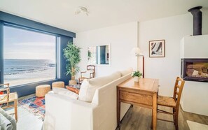 Un saló modern amb un gran finestral amb vista a la platja, una llar de foc i una zona de menjador amb taula i cadires.