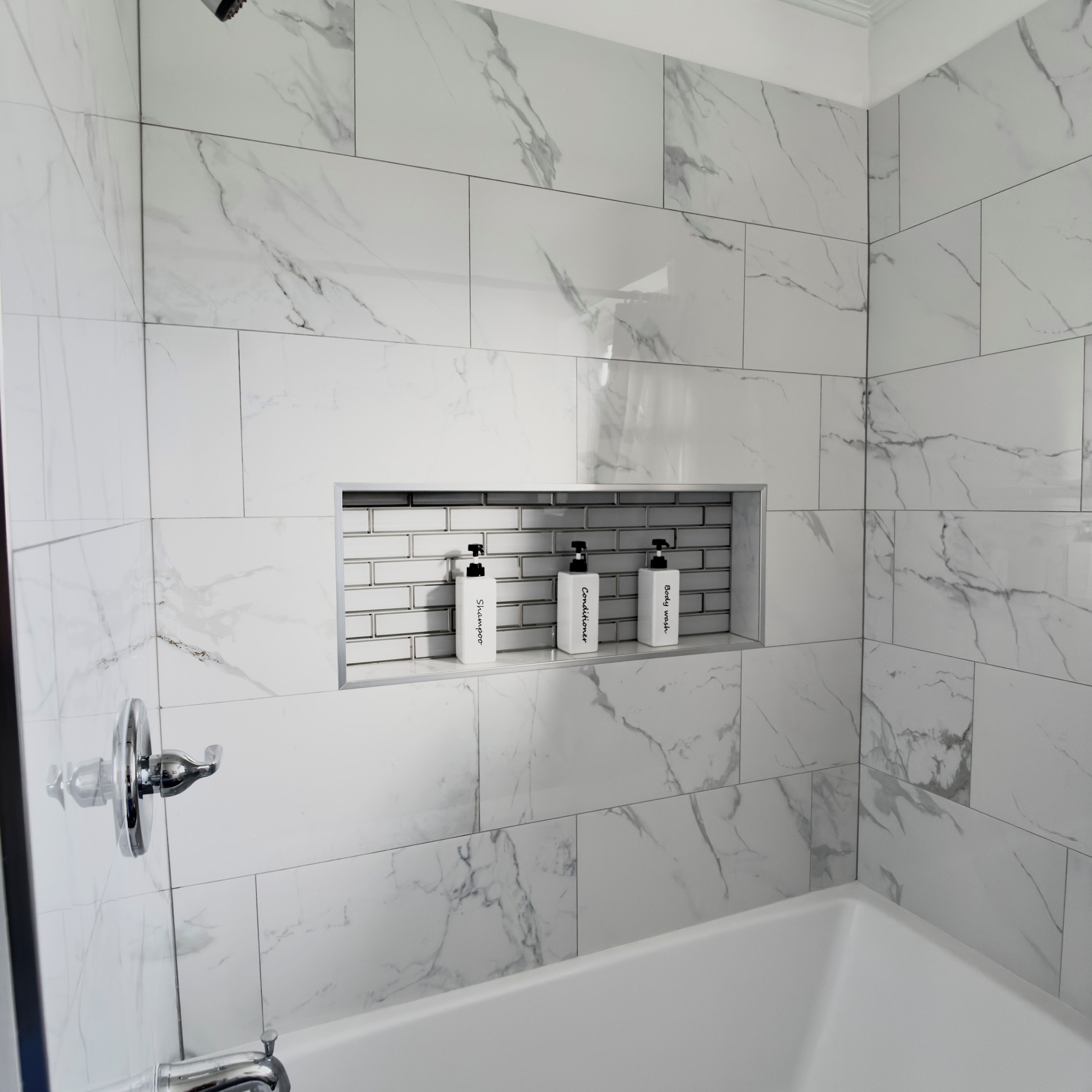 A bathroom with white marble tiles, a built-in shelf with toiletries, and a bathtub.
