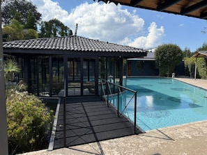A swimming pool with a covered deck area, surrounded by greenery and a clear sky.