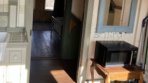 A room with a wooden floor, a microwave on a wooden table, a mirror with a blue frame, and a window with a view of the outside.
