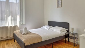 Un lit bien fait, avec du linge de lit blanc et un plaid beige, une table de chevet avec une lampe, et une fenêtre ornée de rideaux légers.