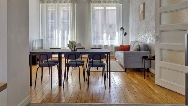 Un salon moderne avec un coin repas, comprenant un canapé, des fauteuils, une table et une grande fenêtre agrémentée de rideaux légers.