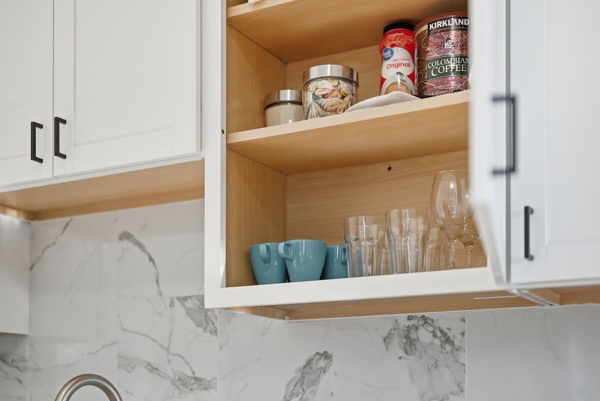 A kitchen cabinet with a marble backsplash, containing a coffee can, a jar, and several glassware items.