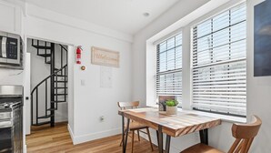 Une cuisine moderne équipée d’un four à micro-ondes encastré, d’une cuisinière et d’une table à manger en bois avec des chaises. On aperçoit un escalier au fond, et un panneau d’accueil est accroché au mur.