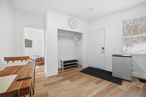 Ein modernes, minimalistisches Zimmer mit Holzboden, einem Bett, einer Bank, einem Schreibtisch, einem Kleiderschrank, einem kleinen Kühlschrank und einer Wanduhr.