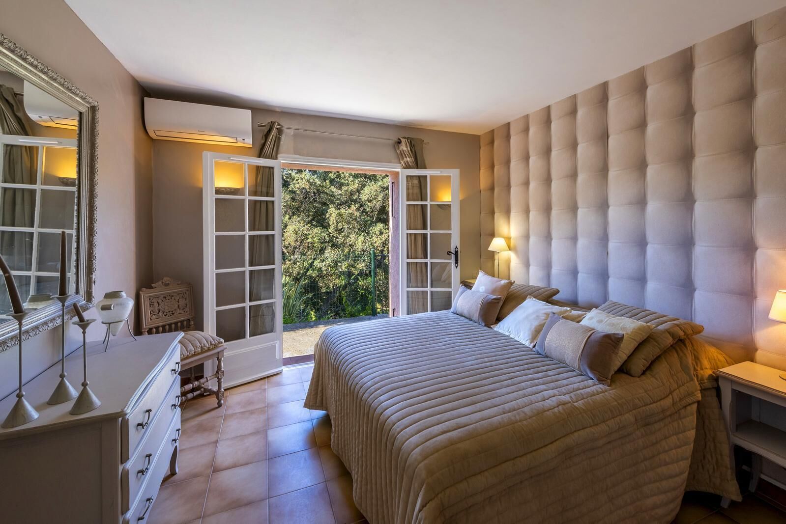 A bedroom with a large bed, a dresser, a mirror, and a window with a view of greenery.