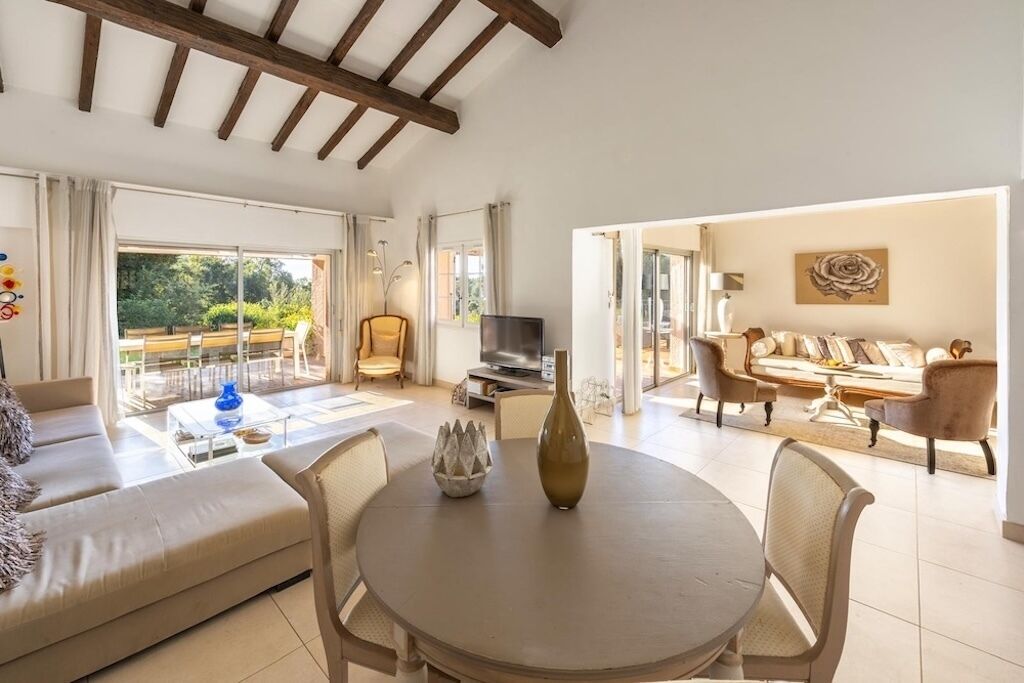 A spacious living area with a sofa, a round table, and a television. There is a balcony with a view of greenery.