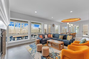 A spacious living room with a sectional sofa, ottomans, and a coffee table. Large windows offer a view of the outdoors. - Sundrift Escape ~ 6BR Luxury Retreat ~ Hot Tub/Sauna/Game Room/Arcade/Golf Cart (Lake Harmony)