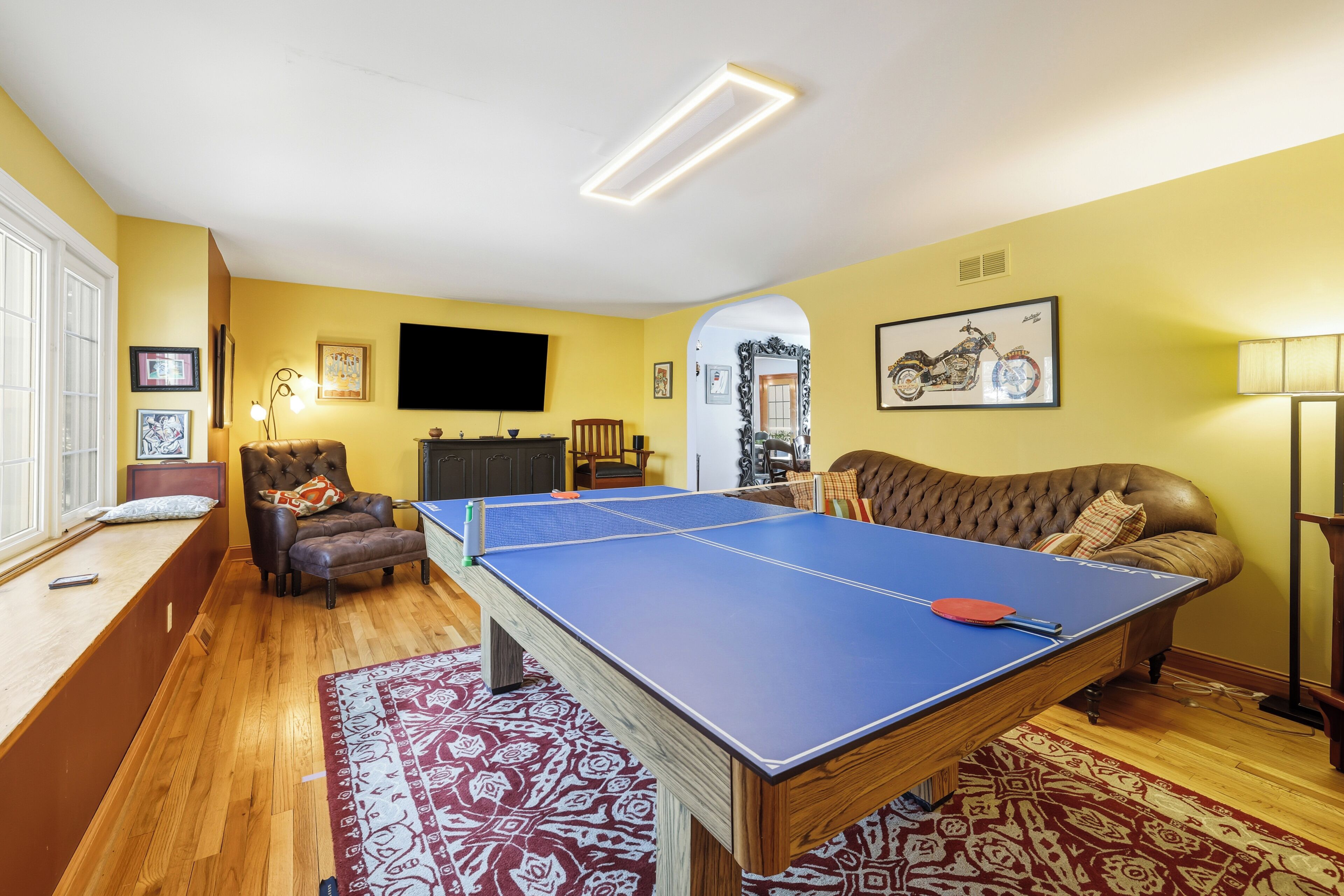 Una habitación con mesa de ping pong, sofá de cuero, televisor y un cuadro de motocicleta en la pared.