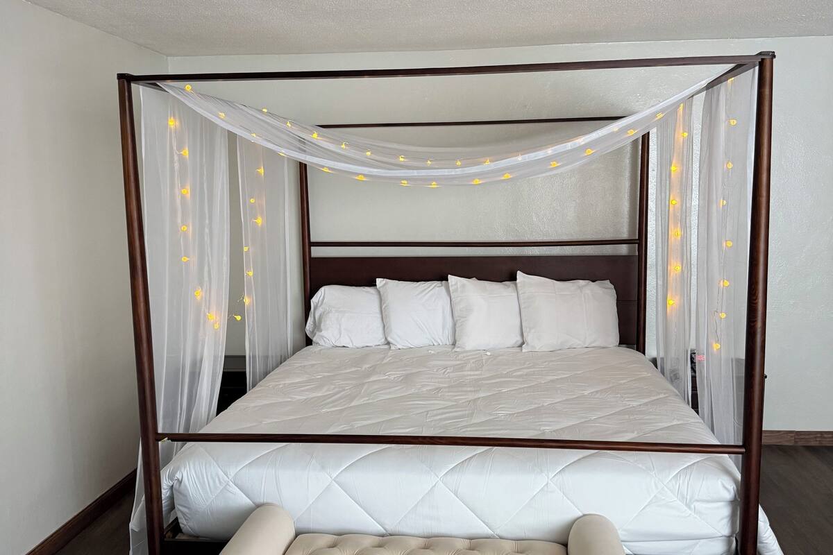 A four-poster bed with a canopy, white bedding, and a tufted bench at the foot.