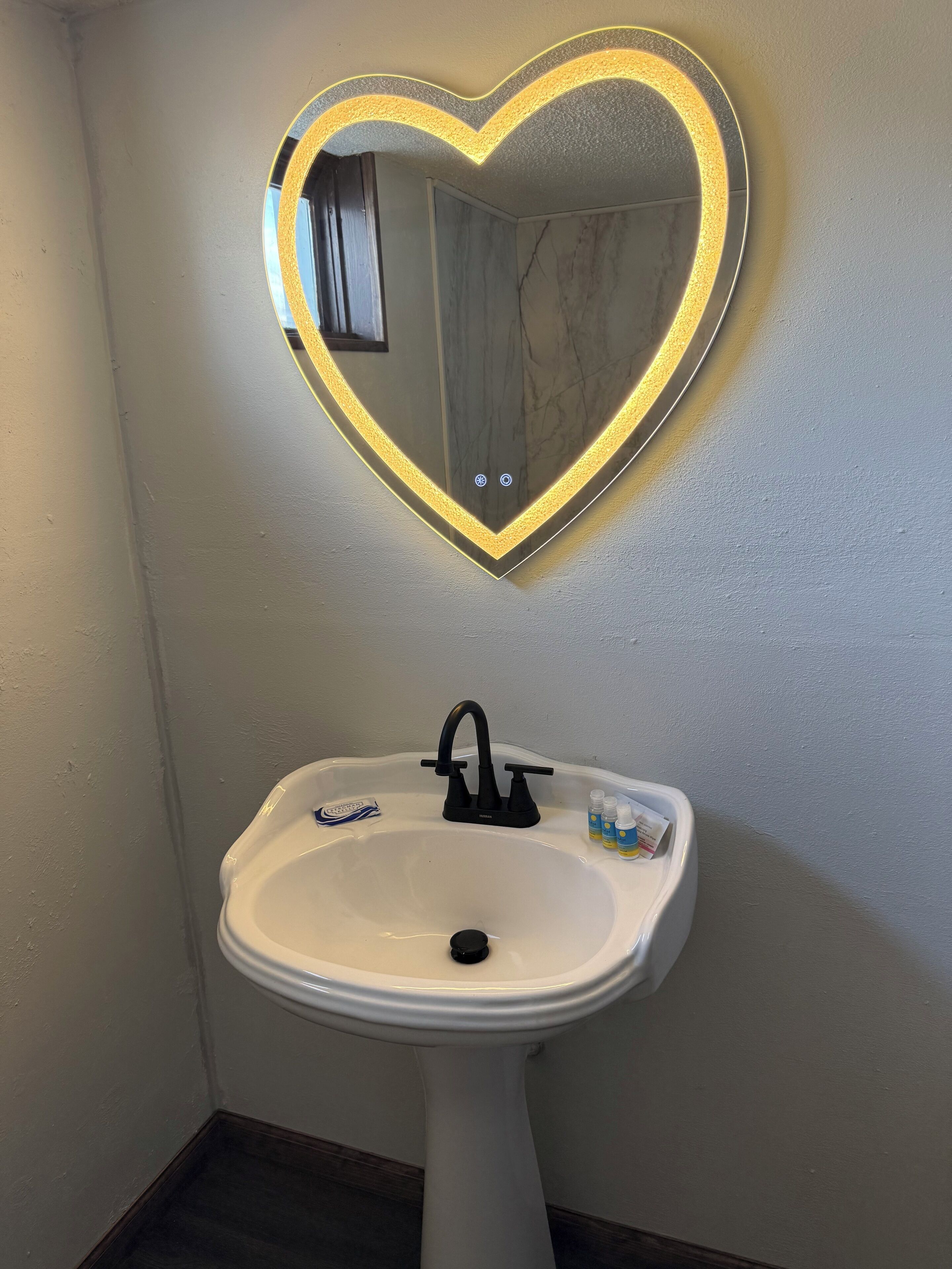 Une salle de bain avec un lavabo sur pied blanc, un robinet noir et un miroir en forme de cœur doté d’un éclairage à LED.
