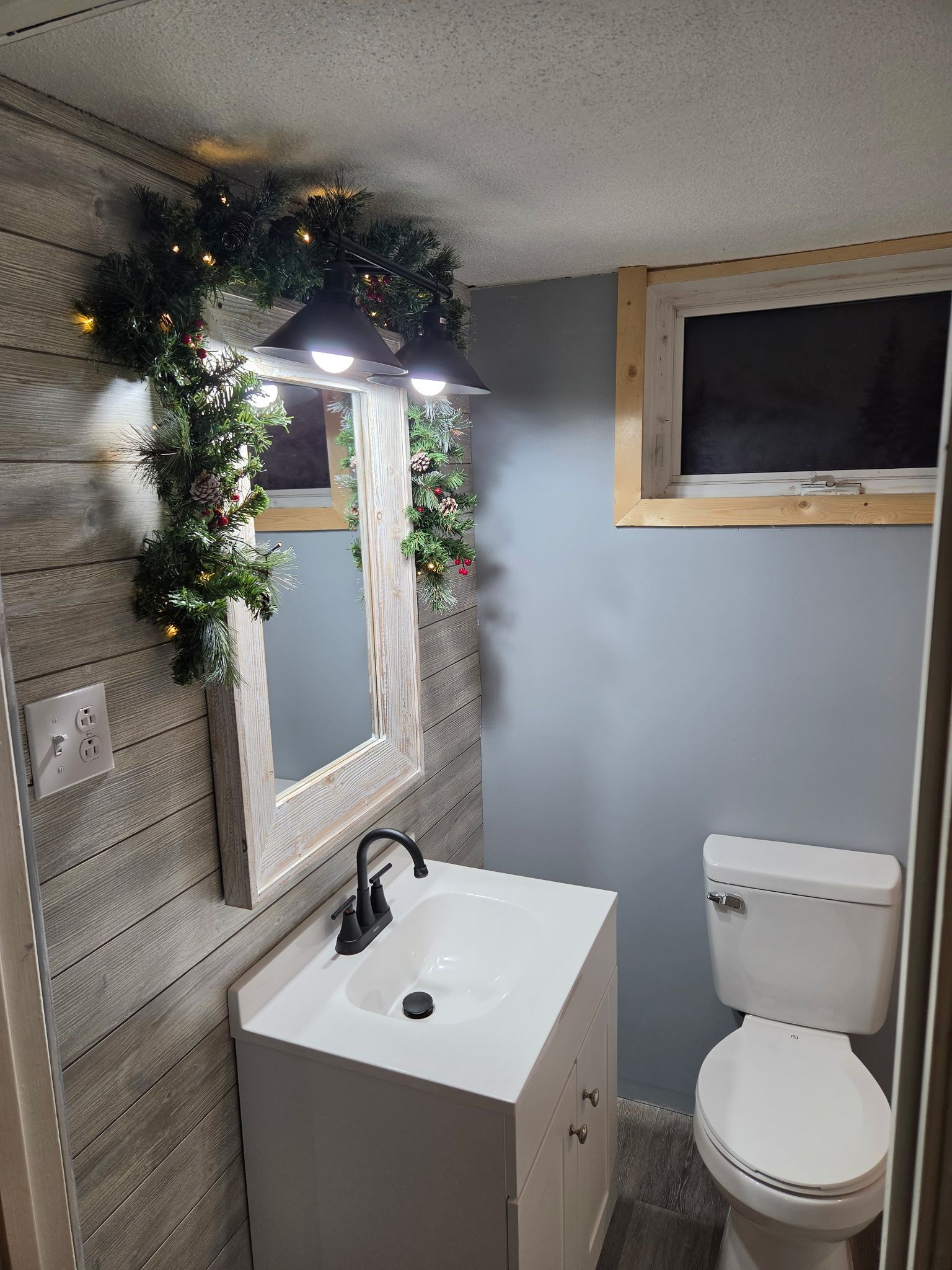 Un baño con un lavamanos blanco, un espejo y un inodoro. Se aprecia panel de pared de madera y una ventana sobre el inodoro.