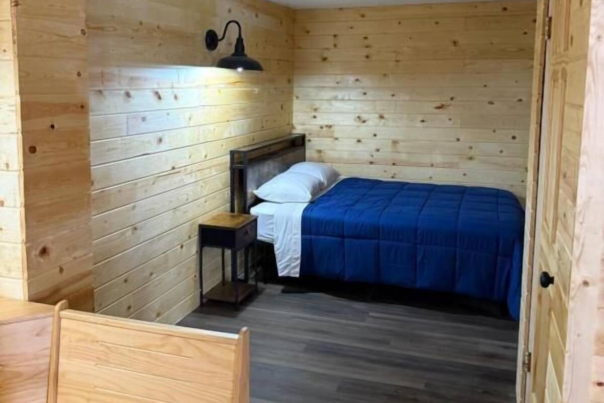 A wooden cabin room with a bed, a small table, and a wall-mounted lamp.