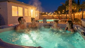 Un grupo de personas en un jacuzzi nocturno, rodeado por una piscina y palmeras, con luces colgantes al fondo.