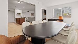 Un interior moderno con una gran mesa ovalada, sillas color beige, una zona de cocina y un ventanal con persianas.