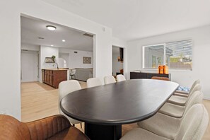 Ein modernes Interieur mit einem großen, ovalen Tisch, beigen Stühlen, einer Küchenzeile und einem Fenster mit Jalousien.