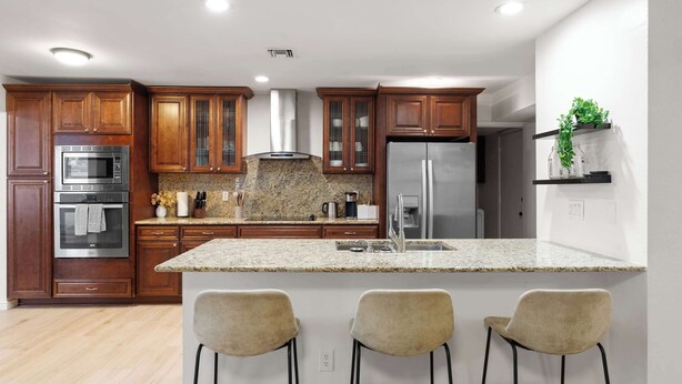 Una cocina moderna con armarios de madera, electrodomésticos de acero inoxidable, encimeras de granito y una zona de bar con dos taburetes.
