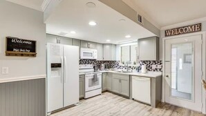 Een moderne keuken met witte kasten, een inbouwoven, magnetron en koelkast. Een welkomstbord hangt aan de muur, en de vloer is van hout.