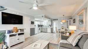 Een moderne woonkamer met een flatscreen televisie, een salontafel en een bank. In de achtergrond bevindt zich een keuken met witte kasten en een eethoek met een tafel en stoelen.